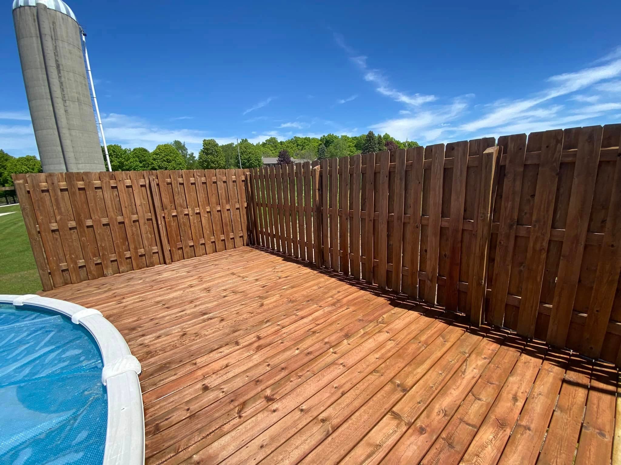 Wooden deck surrounding a pool with blue water, under a clear blue sky and green landscape.