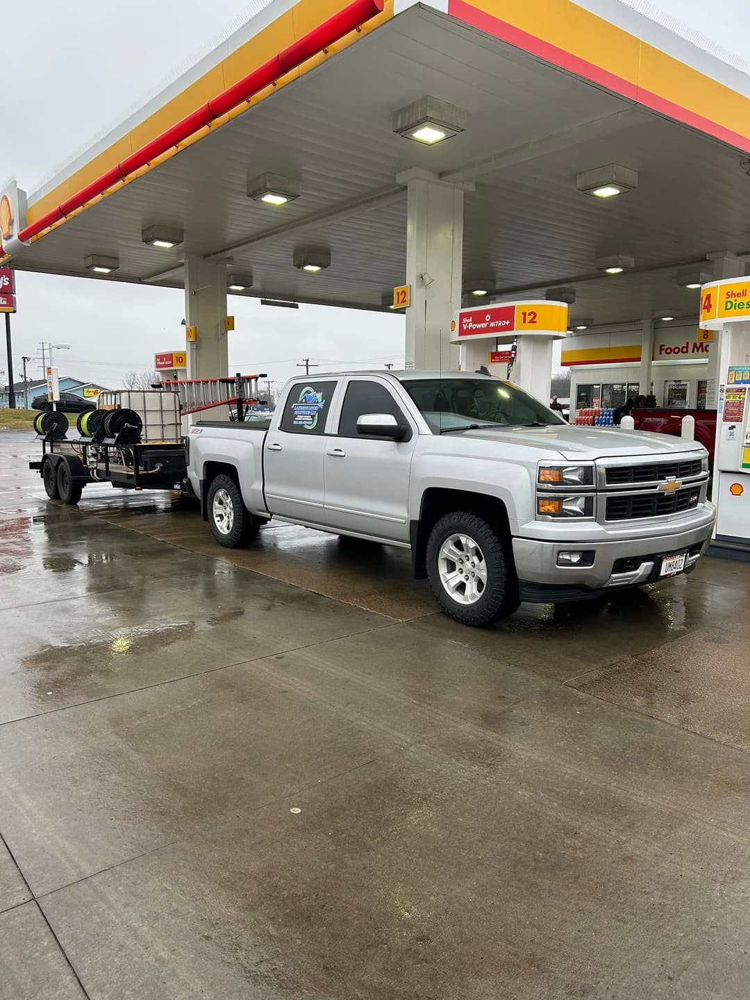 Silver Chevrolet truck with trailer at Shell gas station on a rainy day.