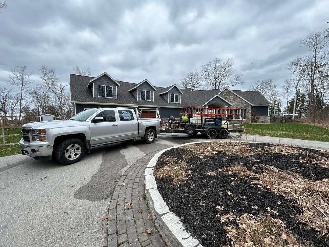 Silver pickup truck towing a trailer parked in front of a large two-story house on a cloudy day.