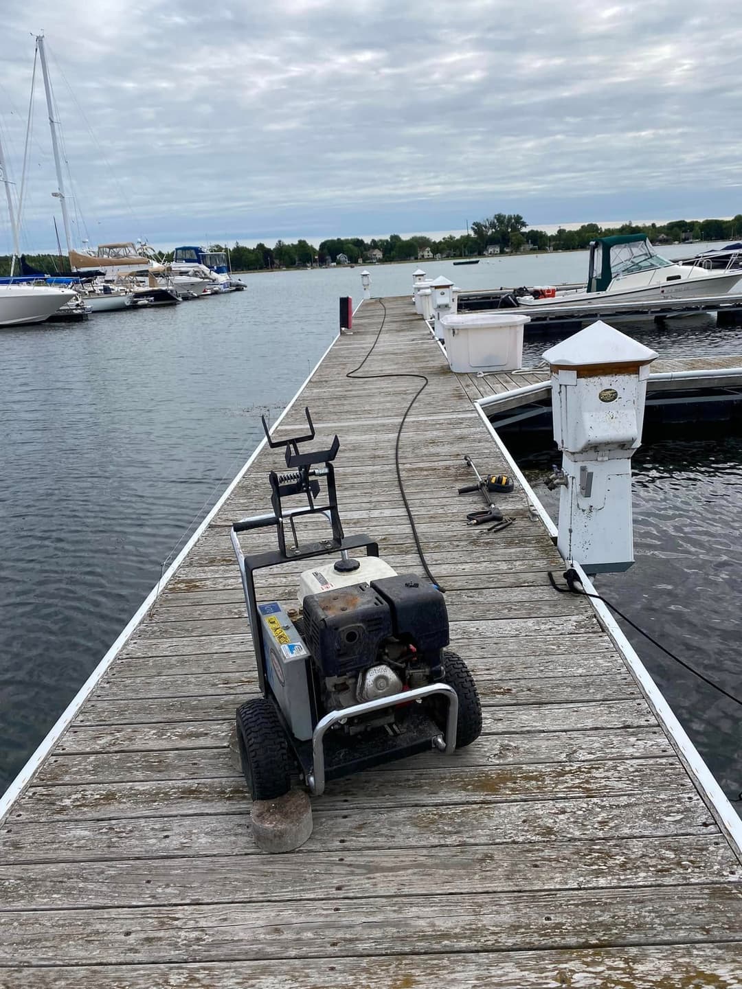 Power washer on a wooden dock near boats on calm water under a cloudy sky.